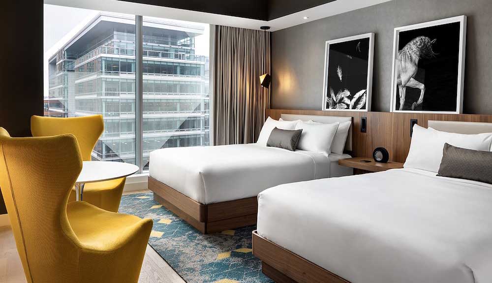 Two queen sized beds are side by side in a room with crisp white sheets and pillows on each bed. There is a decorative grey pillow on each bed. There is a light wood headboard that spans the length of the room and around each bed. There are two mid-century modern style sunshine yellow chairs at the foot of the bed with a round white table between them. From the open window is a high rise building.