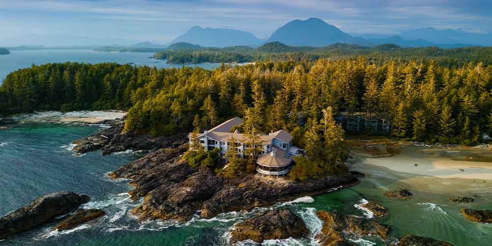 A remote resort with three or four storeys, and a circular area in the front. It is perched atop a rocky area and surrounded by trees behind it, where there is another building that his hidden in the trees. Around the rocky area is turquoise water.