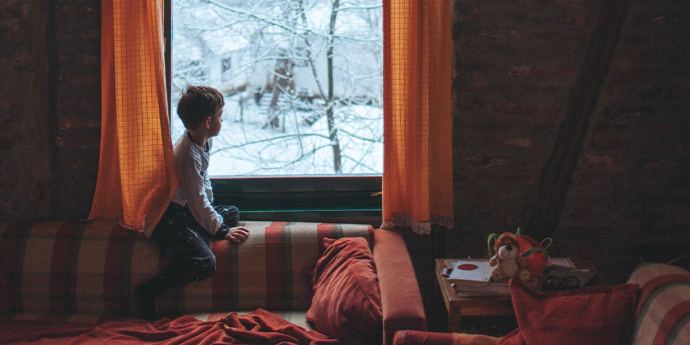 A school-aged boy is sitting on the back of a couch looking out the window. There is another building off in the distance, as well as snow-covered tree branches in front of him. The room he’s sitting in has orange checkered curtains, as well as a rust-coloured couch with a matching pillow on one arm and a matching blanket strewn across teh couch. 