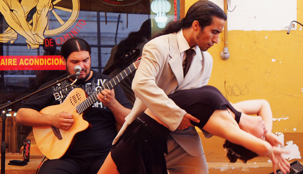 A couple tangoes in front of a live singer playing the guitar, the handsome man with long hair in a ponytail dipping the woman in a black tank top and skirt