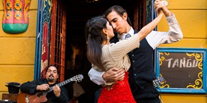 A couple dances the tango in front of a live gutar player sitting in front of a bright yellow building, a sign to the right of the door reads Tango