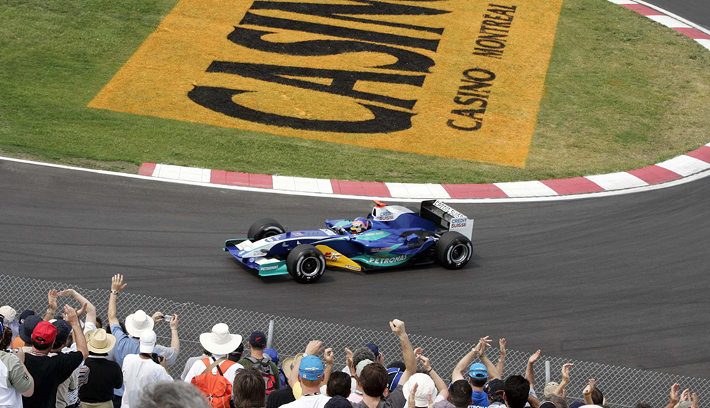 An excited crowd cheer from the stands as a blue race car zips by a curve in the track at the Canadian Grand Prix