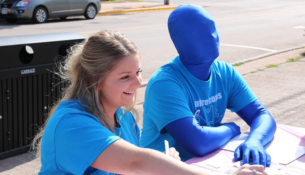 Blonde woman sits next to a man wearing a full blue body suit that hides his face