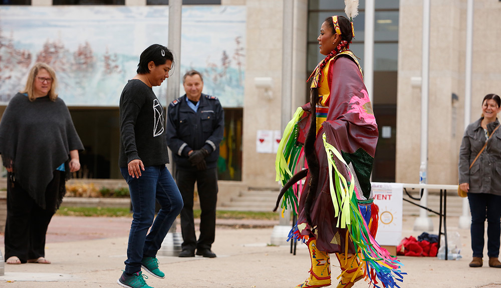 Man learns to dance from a woman dressed in traditional Indigenous wear