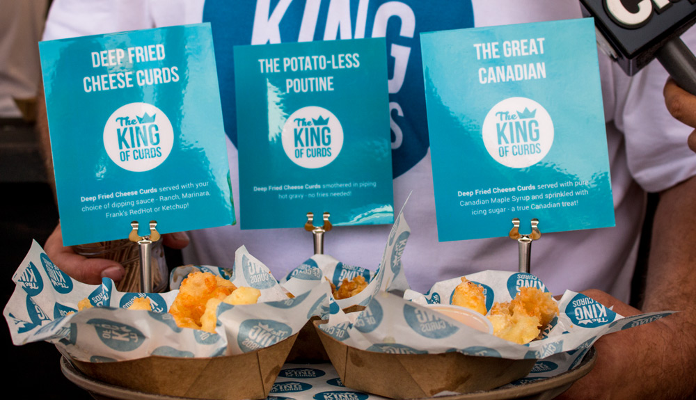 Takeout trays with fried cheese curds, a blue sign atop them reads King of Curds, Deep Fried Cheese Curds, as seen at the CNE