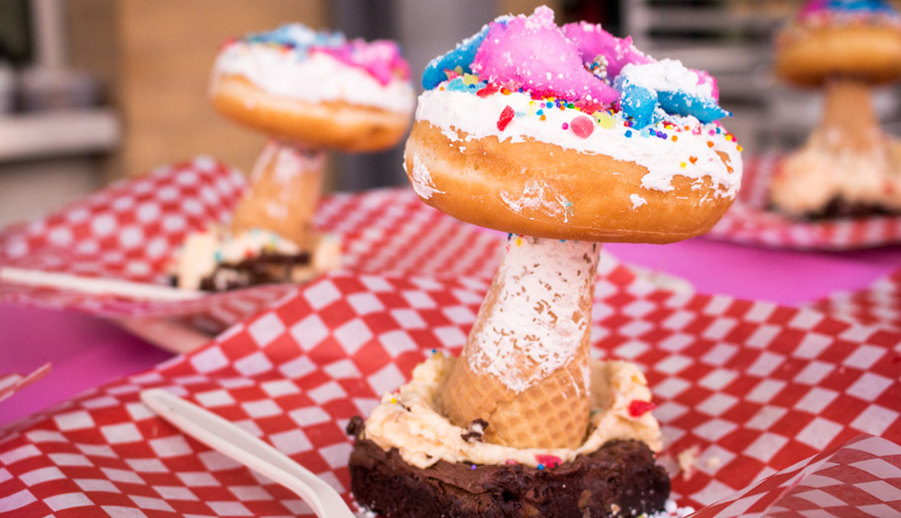 A brownie topped with a sugar cone topped with an iced donut sprinkled with colourful sprinkles and dinosaur-shaped iced cookies, as seen at the CNE