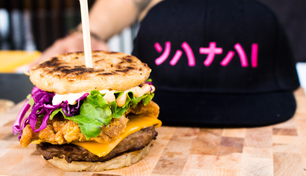 A burger patty is topped with a slice of American cheese, fried chicken katsu, lettuce, purple cabbage and a generous helping of sauce and sandwiched between two buns made of fried ramen noodles, as seen at the CNE