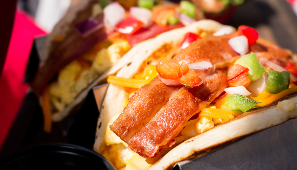 Breakfast tacos made with a pancake shell and stuffed with eggs, melted cheese, green onions, tomatoes, and a slice of crispy bacon, as seen at the CNE