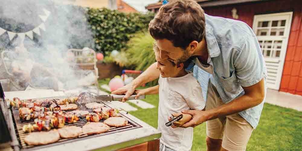 A man in a blue button-up shirt and beige shorts standing behind a child in a white T-shirt who are both holding onto a pair of tongs in front of a smoky barbecue, which has some burger patties on it as well as meat and vegetable kebabs.