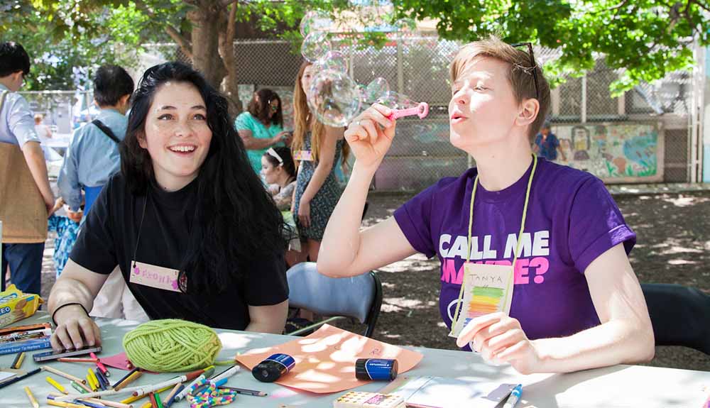 Two young people sit behind a table outdoors with craft materials on it, one blowing a large bubble
