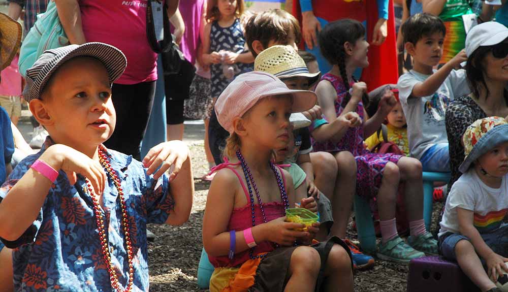 A crowd of young children and their parents sit watching a show, the children wear multi-coloured beads and event wristbands