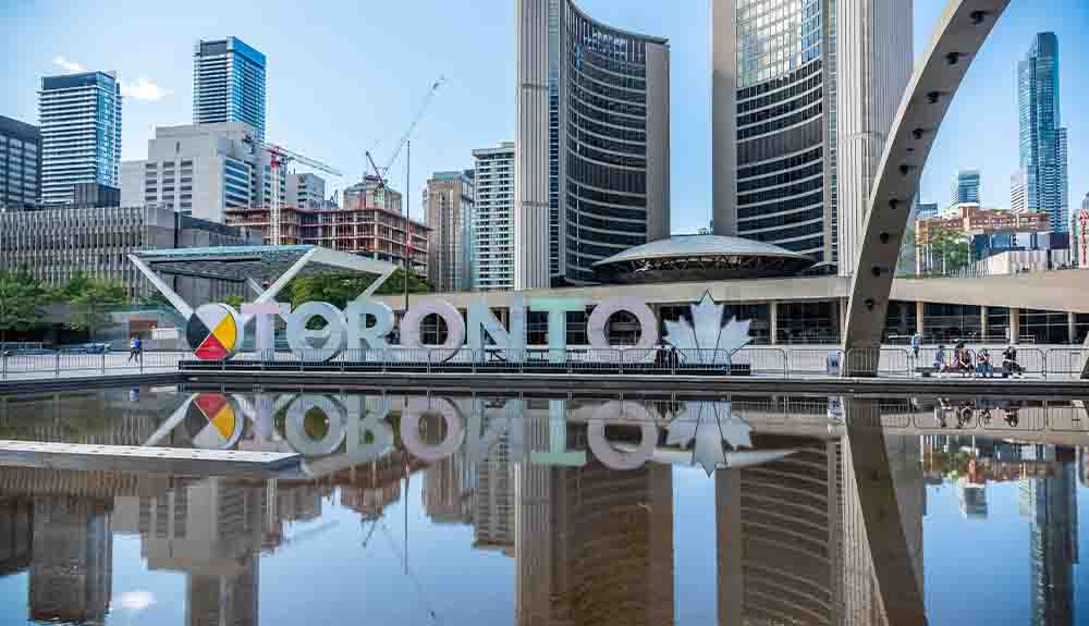 Nathan Phillips Square in Toronto with the Toronto sign lit up in red, blue and grey with City Hall pictured in the back. There are two tall office towers that are curved in with a smaller dome at ground level.