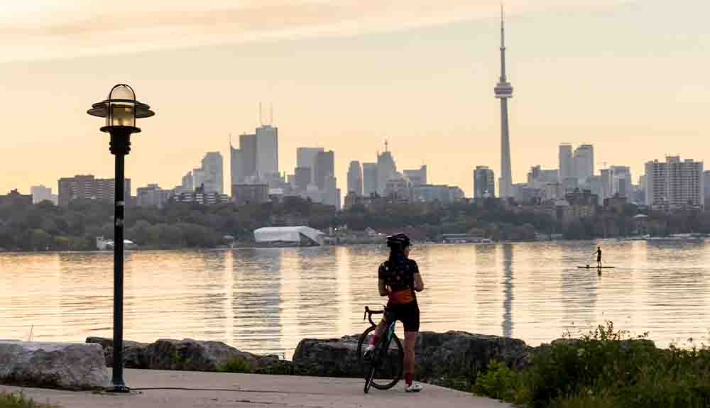 A cyclist is straddling her bike and looking at Lake Ontario back at the city of Toronto. The CN Tower can be seen in the skyline alongside other office towers and condo buildings.
