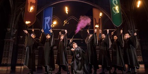 The cast of Harry Potter and the Cursed Childs stands wearing black cloaks and holding wands up in the air. Some shoot small bursts of fire, while the centre person has pink mist coming from his wand. Red, blue, orange and green banners of Hogwarts houses are seen hanging in the background.
