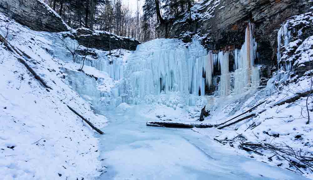 A close up of a frozen waterfall with cascades of frozen blue and white water hanging from brown rock formations. To the left side, there is a steep hill covered in snow.