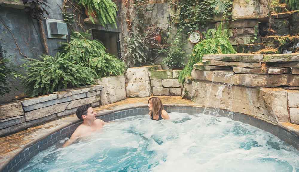 A man and a woman are sitting in a circular pool. Behind them there is a small waterfall with water flowing into the pool they're in. There is a door in the corner that is surrounded by lots of greenery. 