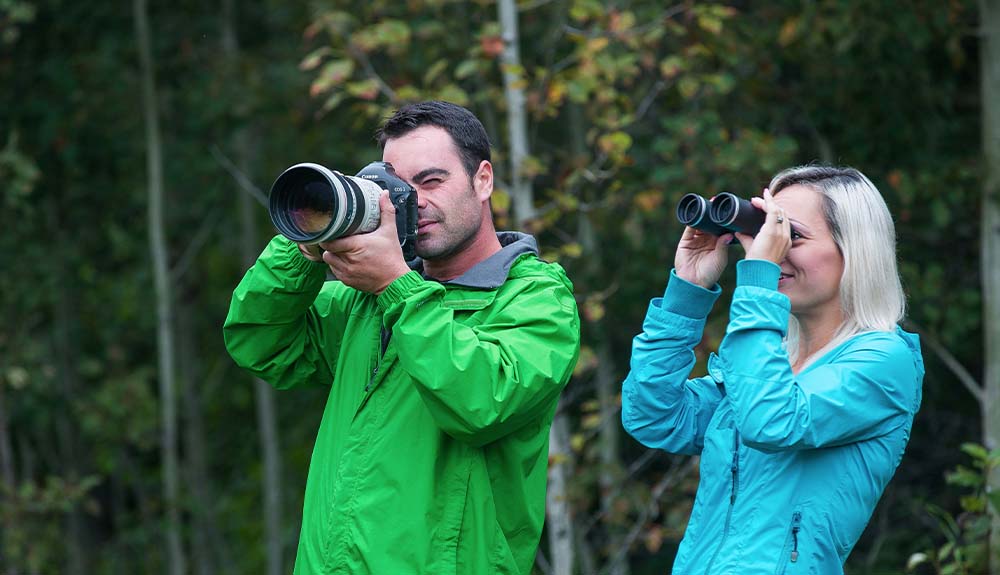 Two people are standing next to each other in the woods. There is a man wearing a bright green coat with a grey collar over the coat. He is holding a Canon SLR camera with a very wide lens. Next to him is a woman with white-blonde hair wearing a sky blue coat. She is holding a pair of binoculars and looking off into the distance.