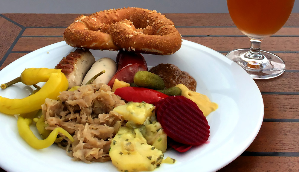 Sausages, saurekraut, potato salad and pickled vegetables fill a plate with a pretzel on the side