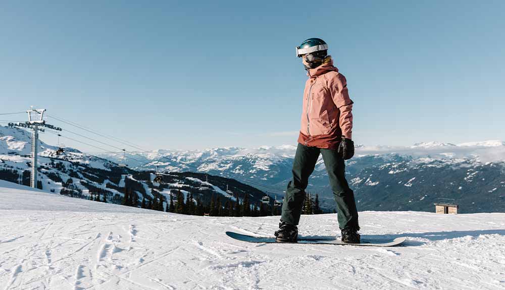 A person wearing a burnt orange ski jacket and black snow pants standing on top of a snowy hill, looking out at the snowcapped mountains around them. They are wearing a black helmet with goggles on top of the helmet and standing on a snowboard.