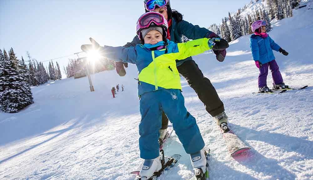 A person wearing a teal ski jacket and black snow pants is standing behind a child who is wearing a teal and neon yellow ski jacket and ski pants. The child has ski goggles on over their forehead. Both people are on skis but do not have poles. The child is holding their arms out for balance.