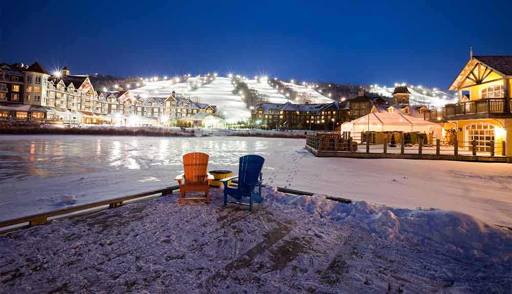 Two Muskoka chairs, one red and one blue, are sitting on a dock. There is a pathway of snow behind it. Even further behind it is a cluster of buildings on a hill that are brightly lit up. Behind the buildings are downhill ski slopes that are well-lit for night skiing.