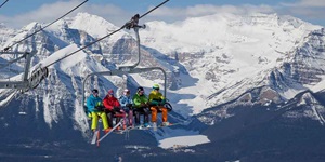 Five people are sitting on a ski lift with snow-capped mountains behind them. The first person is wearing a black helmet with a teal coat and neon yellow snow pants. The second person next to them is wearing a red coat, black snow pants and a red helmet. The person in the middle is wearing a checkered powder blue coat with pink snow pants and a white helmet. The fourth person is wearing a neon green coat with black ski pants and a black helmet. The last person is wearing a multi-coloured coat of neon yellow, teal and black with a matching helmet, and orange snow pants.