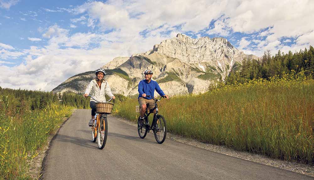 A woman and man wearing helmets enjoying a bike ride along a paved pathway on the Banff Legacy Trail