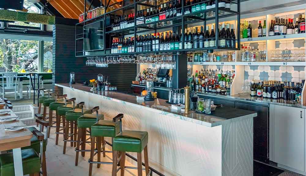 A view of a restaurant and a bar space with green leather chairs, a long island counter with a white counter top and wine bottles sitting on shelves over the island. Behind the counter is a blue and white tiled wall with more wine bottles and other alcohol bottles.