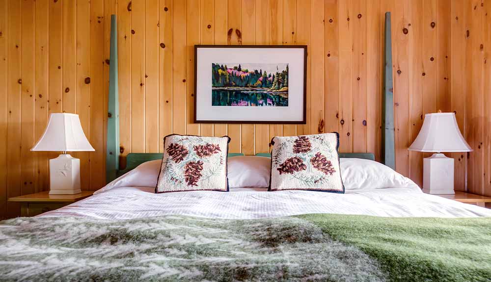 A bedroom with pine plank walls and a wooden bed frame painted in light green. There are white lamps on night stands flanking the bed, which has light pink sheets and two decorative throw pillows with pinecones on it.