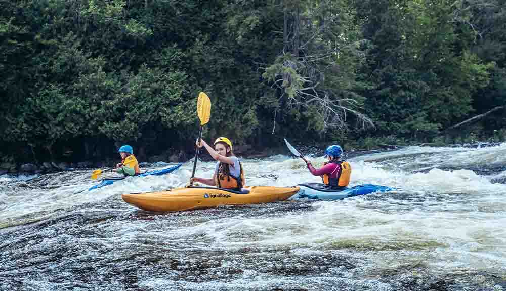 Three young people are paddling in yellow kayaks. They are each wearing helmets on gentle rapids with dark green trees behind them.