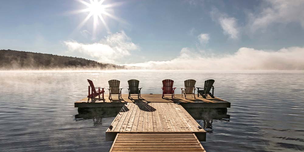 A wooden dock on the water with six Muskoka chairs sitting at the end of it, facing the lake and a cloudless sky and the sun shining down on it.