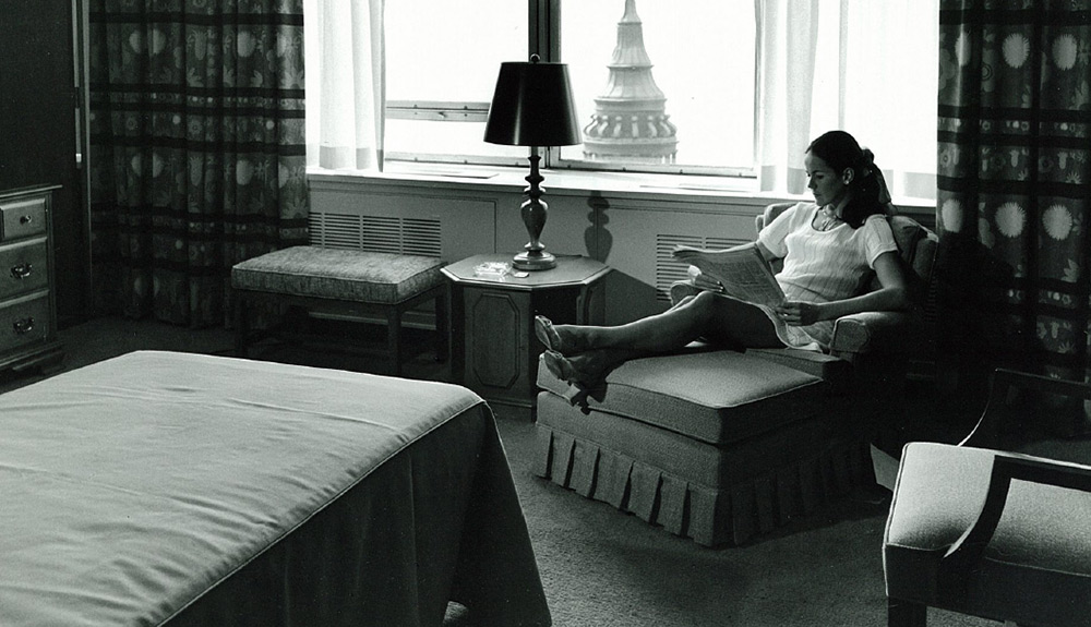 Black and white photo of a woman reading the paper in armchair with her legs up on an ottoman