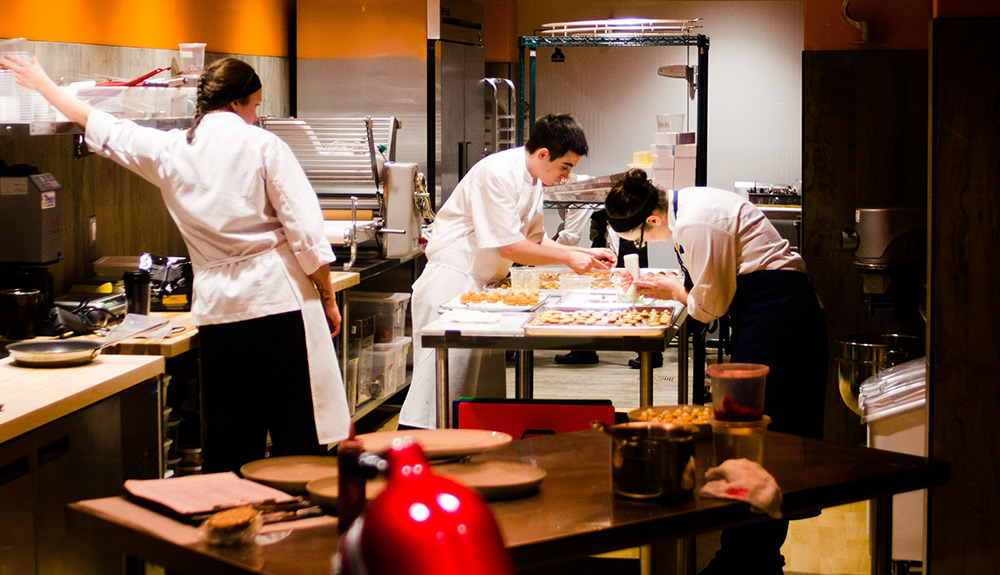 One female chef reaches for something on a shelf as two other chefs work on decorating pastries