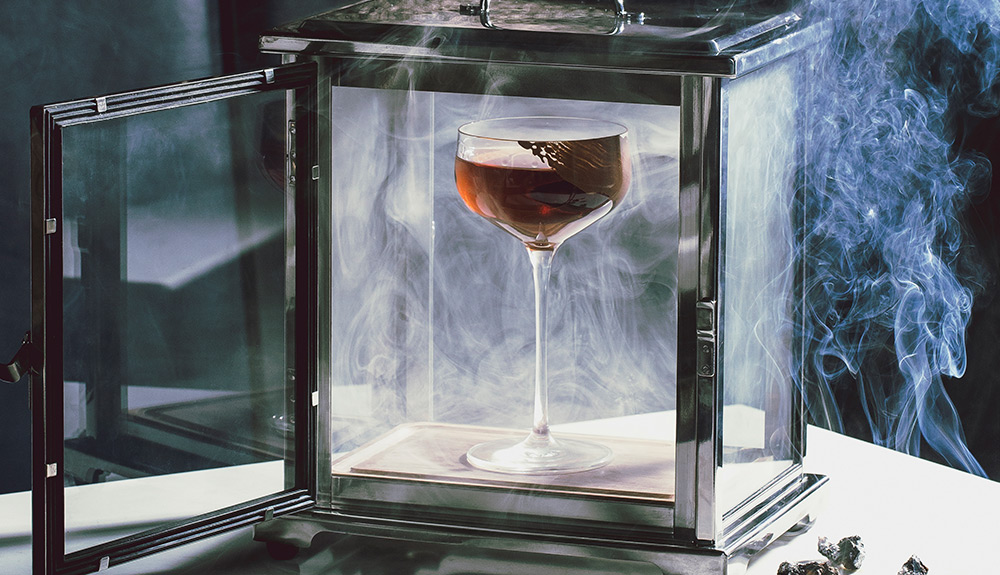 A fancy cocktail is presented in a glass box with smoke swirling around it