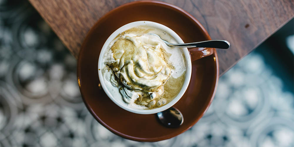A round mug with a spoon inserted, filled with coffee and white gelato sits on top of a brown saucer with a second spoon. The image for this Italian affogato is taken from above. It sits on the corner of a wood table on a blurred black and white, circular-patterned floor. 