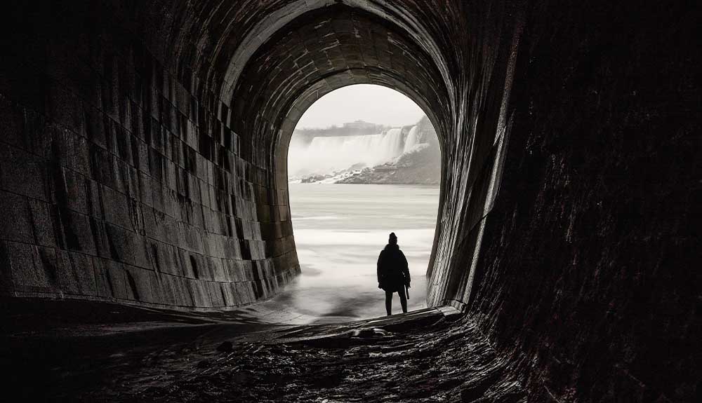 A lone person with his back to the camera standing under a curved stone arch looking at Niagara Falls.