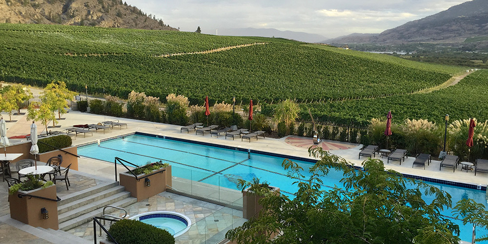 A luxury resort with outdoor hot tub and long pool are seen beside the hilly vineyards in Okanagan