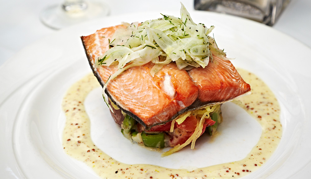 Fine dining plate of a vegetable salad, salmon fillet with skin on and cucumber salad on top, plated with a Dijon mustard sauce