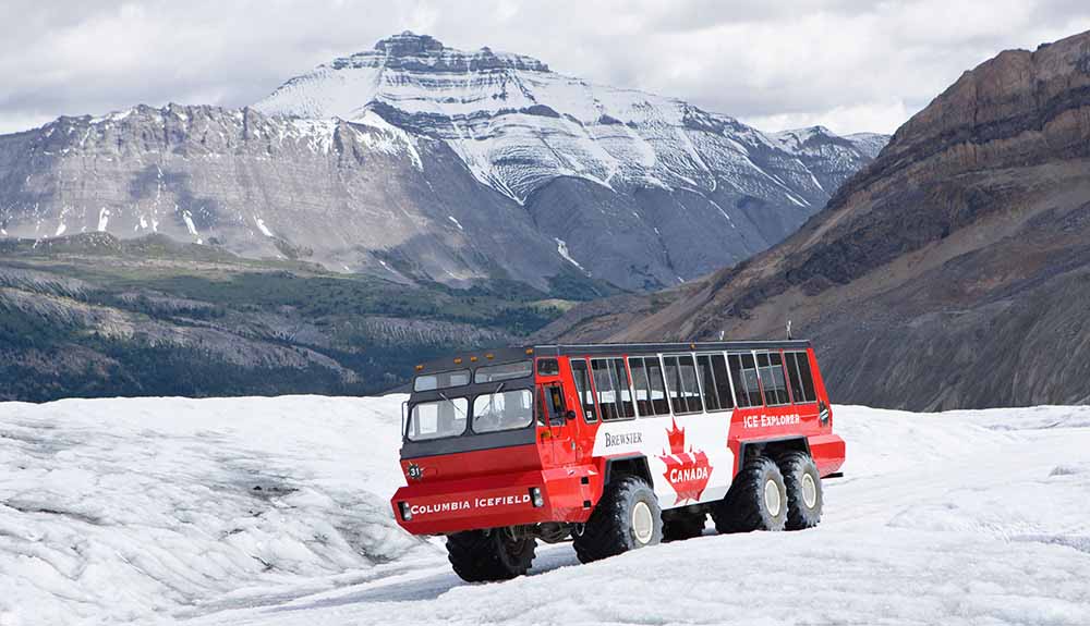 A red bus with large all-terrain wheels and maple leaf logo on the side by the ice fields in front of the Canadian Rockies