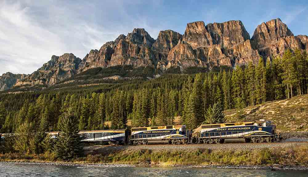 Rocky Mountaineer train drives along the rail track by the water in front of the Rocky Mountains