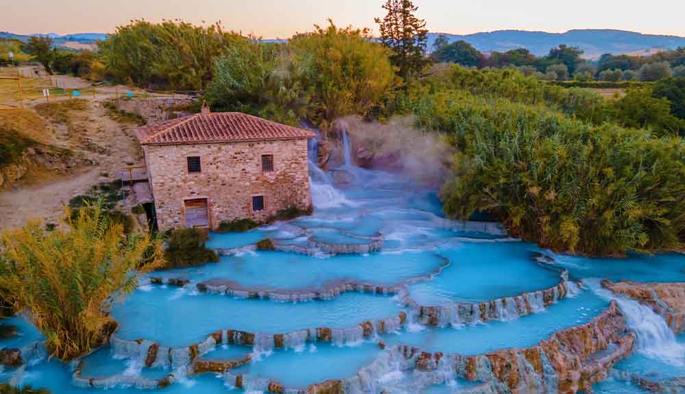 Cascate Del Mulino in Tuscany Italy. Aqua-marine water flows downward from pool to pool across a rocky hillside. A brown brick house is seen at the top of the hill and lush green forest in the distance. 