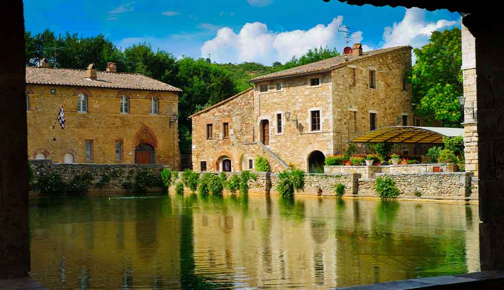 Bagno Vignoni in Tuscany, Italy. Green water in the middle of a town piazza, with light brown brick buildings surrounding. There is green foliage in the brick wall around the body of water and behind the buildings. The sky is blue with white fluffy clouds.