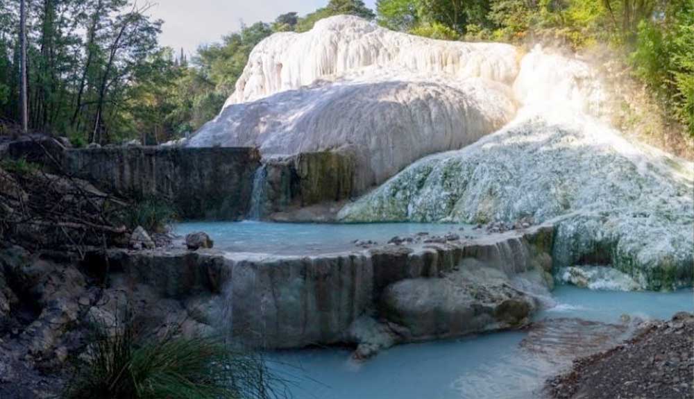 Fosso Bianco in Tuscany, Italy. A large white calcium formation sits on a hill with two light blue pools of water that below span the rocky hillside. A few bathers wade in the waters. There is green foliage seen to the left and behind. 