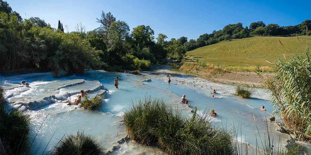 Aquamarine-coloured thermal pools sit across a tapered hillside. There are small waterfalls flooding downward from pool to pool. A brown brick house sits on the top of the hill in the centre, which has a dirt road leading to it from the left. The area is surrounded by green vegetation.