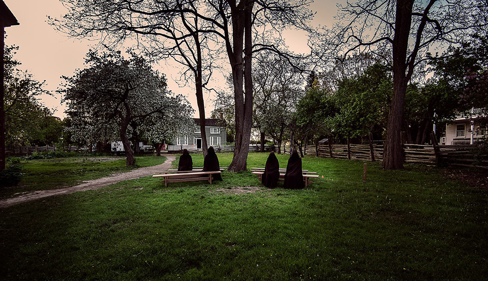 Four cloaked people sit on benches by naked trees in the fall
