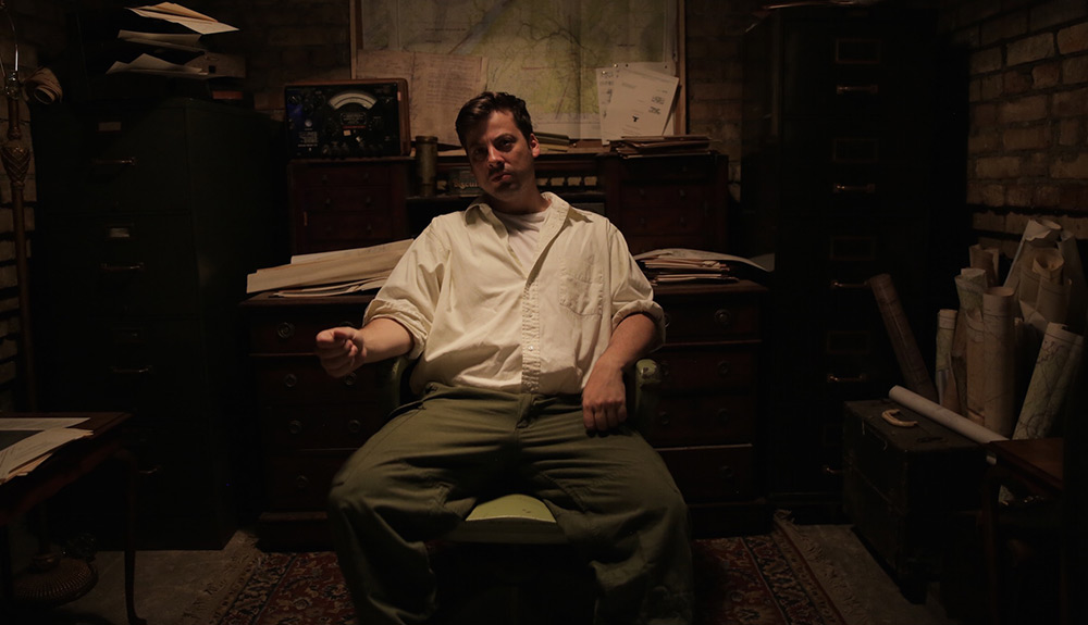 Man wearing white button up shirt and hunter green slacks sits in a chair