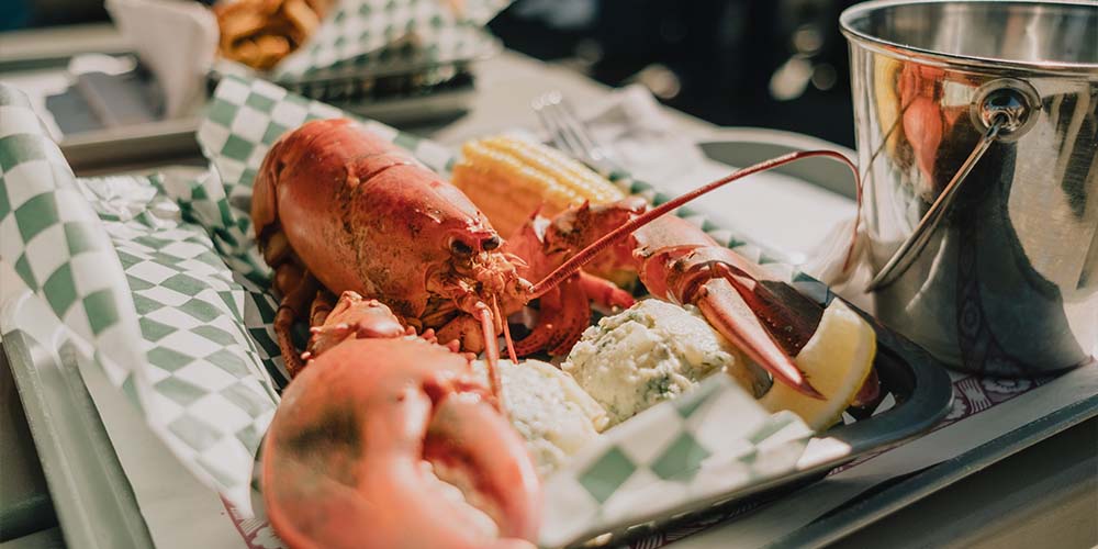 A cooked red lobster sits with mashed potatoes, a small cob of yellow corn and a lemon on top of a black tray lined with a green and white checkered napkin. There is a small silver bucket seen to the right and a fork behind it. In the background you see another blurred tray of food.