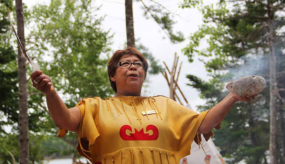 A woman wearing a yellow shirt with a red design on the front holds a feather in one hand and a bowl in the other hand with some smoke around it performs a smudging ceremony.