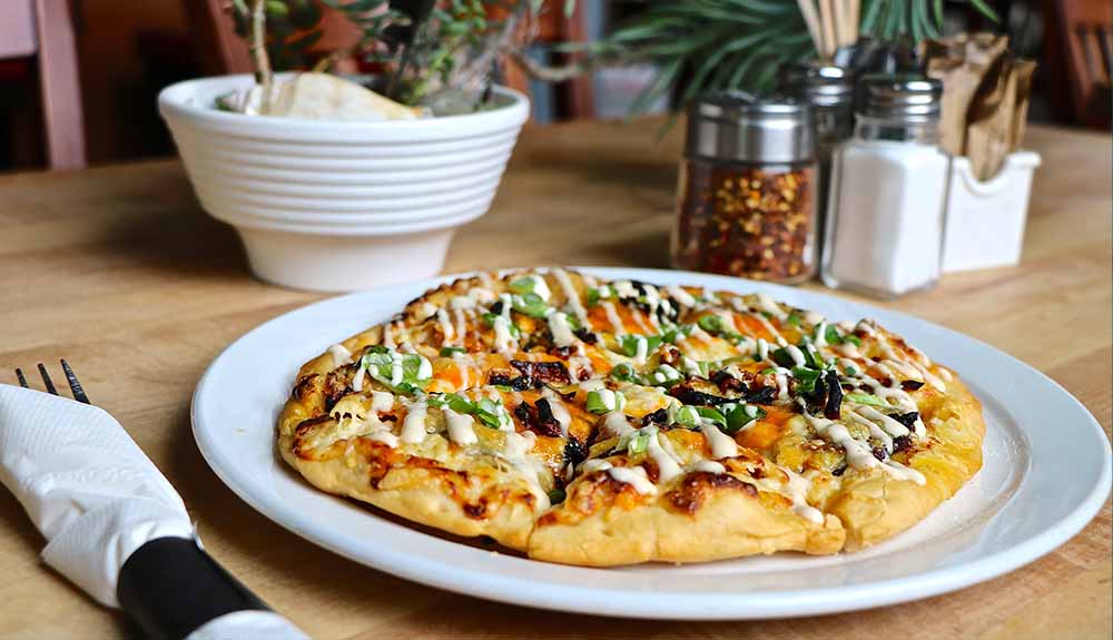 A table set with a fork rolled up in a white tissue next to a plate of bannock pizza, which is topped with green onions and a white sauce. A plant in a white pot and chili flakes can be seen in the background.