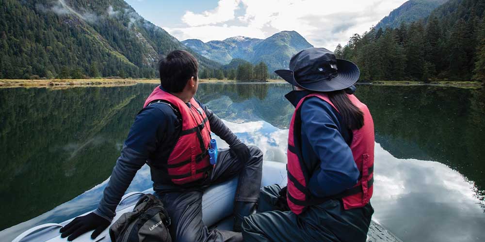 Two people wearing red vests sitting in a boat with their backs to the camera looking at the mountains behind them.